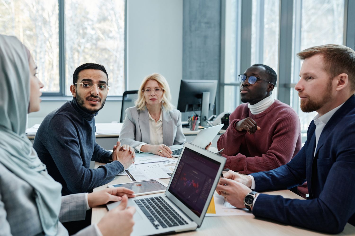 A diverse team talks around a meeting table with laptops.